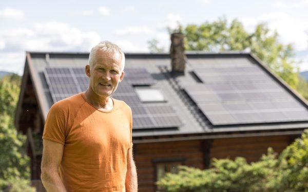 Vegard Ulvang står i oransje t-skjorta foran brunt hus med solcellepanel på taket.